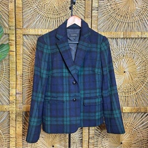 Pode incluir: Blazer xadrez azul marinho e verde com dois botões e dois bolsos frontais. O blazer está pendurado em um cabide de madeira contra um fundo tecido. Um blazer de estilo clássico.