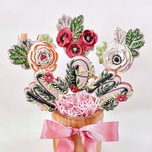 May include: A wooden planter with a pink ribbon bow holds a bouquet of decorated cookies. The cookies are shaped like flowers and the letter "M".