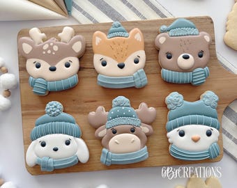 May include: A collection of six winter-themed sugar cookies. Each cookie is shaped like a different animal: a deer, fox, bear, dog, moose, and penguin. They are decorated with blue hats and scarves. The cookies are arranged on a wooden board.