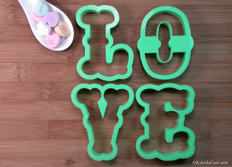 May include: Green plastic cookie cutters shaped like the letters L, O, V, and E. The cutters are on a wooden surface.