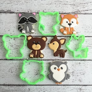 May include: A set of nine cookie cutters featuring various woodland animals, including a raccoon, fox, bear, deer, owl, and three generic animal shapes. The cutters are made of green plastic and are arranged on a white wooden surface.