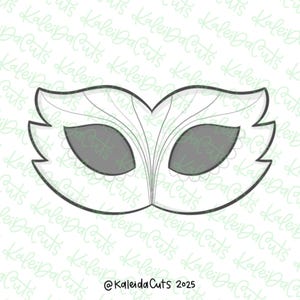 May include: A white mask-shaped cookie cutter with a decorative design. The mask has eye holes and is outlined in black. The text "@KaleidaCuts 2025" is at the bottom.