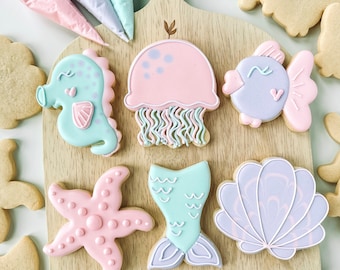 May include: A wooden cutting board with a variety of decorated sugar cookies. The cookies are shaped like a starfish, a seashell, a jellyfish, a mermaid tail, a seahorse, and a fish. The cookies are decorated with pastel colors and icing.