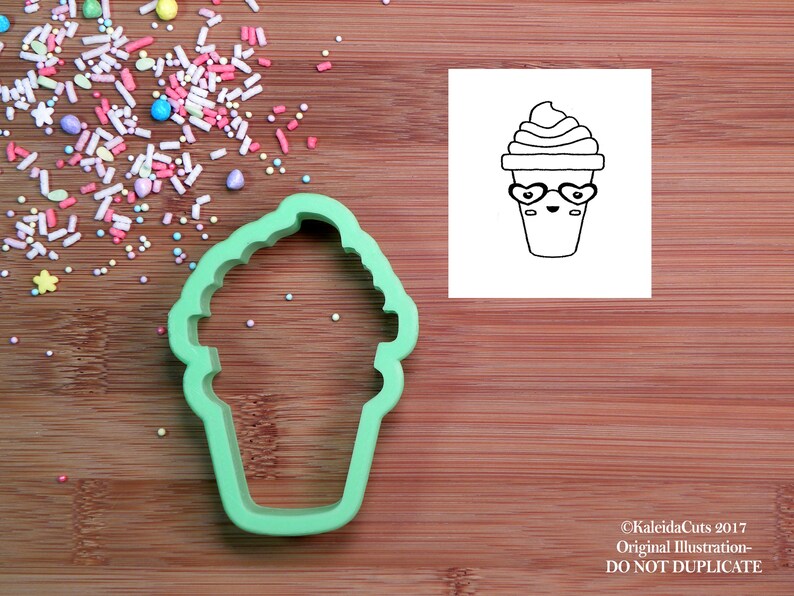 K&ouml;nnte beinhalten: Gr&uuml;ner Plastik-Ausstecher in Form eines Cartoon-Eisbechers mit einem l&auml;chelnden Gesicht und herzf&ouml;rmigen Sonnenbrillen. Der Ausstecher befindet sich auf einer Holzoberfl&auml;che mit bunten Streuseln.