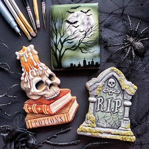 May include: Three Halloween-themed sugar cookies. One cookie is a green and brown scene with a moon and bats. Another cookie is a tombstone with the words "RIP" on it. The third cookie is a skull with a candle on top of it, sitting on a stack of books that say "Potions".