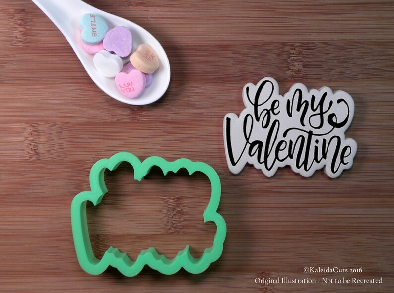 May include: Green plastic cookie cutter shaped like the words "Be My Valentine" on a wooden surface. A white ceramic spoon with colorful candy hearts is in the upper left corner.