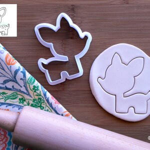 May include: White plastic cookie cutter shaped like a cartoon deer. The cutter is shown on a wooden surface with a rolling pin and a piece of white fondant that has been cut out with the cutter.