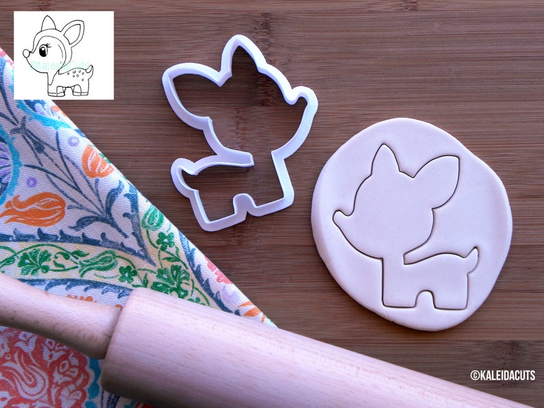 May include: White plastic cookie cutter shaped like a cartoon deer. The cutter is shown on a wooden surface with a rolling pin and a piece of white fondant that has been cut out with the cutter.