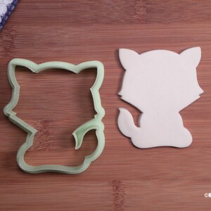 May include: A green plastic cookie cutter shaped like a cartoon cat, next to a white fondant cutout of the same shape.