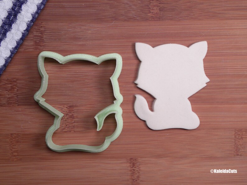 May include: A green plastic cookie cutter shaped like a cartoon cat, next to a white fondant cutout of the same shape.