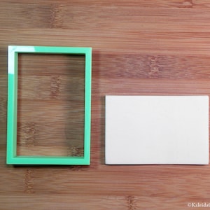May include: A green plastic rectangle cookie cutter in the shape of a frame, and a white rectangular piece of fondant on a wooden surface.
