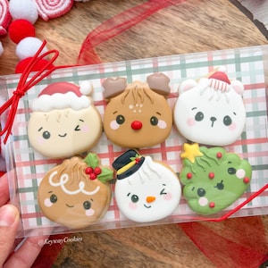 May include: A clear plastic box containing six decorated sugar cookies with holiday themes, including a Santa-hatted face, a reindeer, a polar bear, a gingerbread cookie, a snowman with a top hat, and a Christmas tree, labeled KeywayCookies.