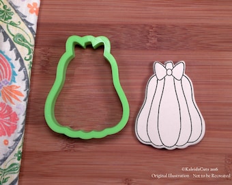 May include: Green plastic cookie cutter shaped like a pumpkin with a bow on top.  Next to it is a white outline of the same pumpkin on a brown wooden surface.