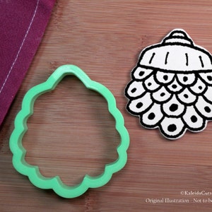 May include: Green plastic cookie cutter shaped like a pine cone. The cookie cutter is on a wooden surface next to a white and black illustration of a pine cone.