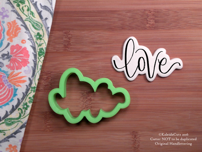 May include: Green plastic cookie cutter shaped like the word "love" in a handwritten style. A white cookie with black icing is shown next to the cutter.