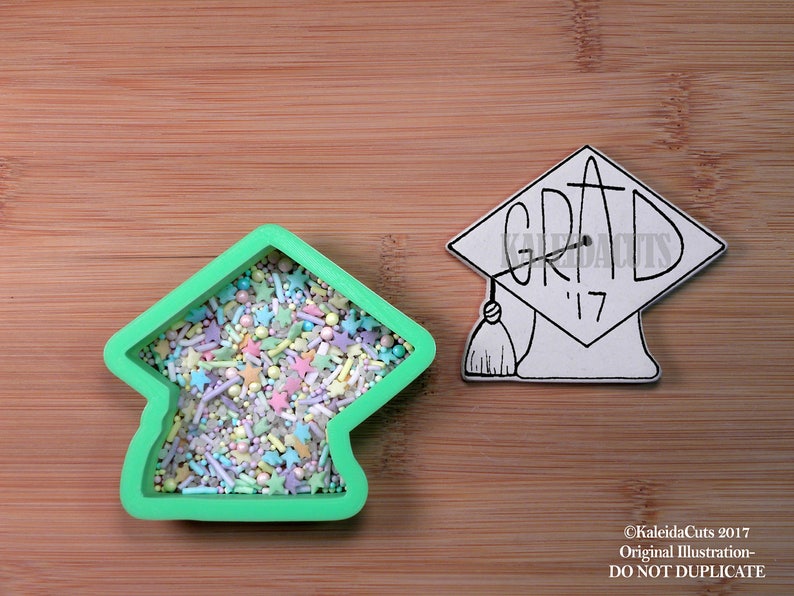 May include: Green plastic cookie cutter shaped like a graduation cap. The cutter is filled with colorful sprinkles. A white graduation cap with the word "GRAD" and the year "'17" is shown to the right of the cutter.