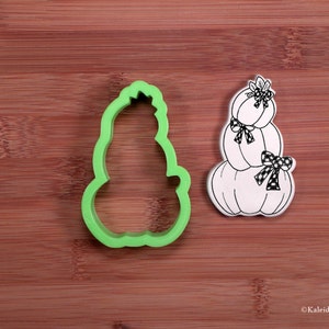 May include: Green plastic cookie cutter shaped like a stack of three pumpkins with a bow on the bottom pumpkin. The cutter is next to a black and white illustration of the same design.