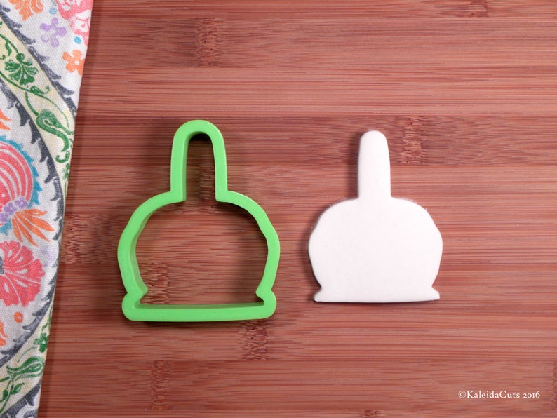 Op de afbeelding: Groene plastic koekjesvormer in de vorm van een kerstboomversiering. De vormer ligt op een houten oppervlak naast een witte plastic koekjesvormer van dezelfde vorm.