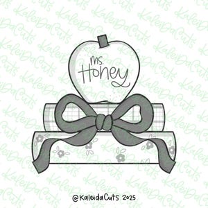 May include: A grayscale illustration of an apple with the text "ms. Honey" above a stack of books tied with a bow. The books have floral and plaid patterns. The design is likely for a teacher or school-related theme.