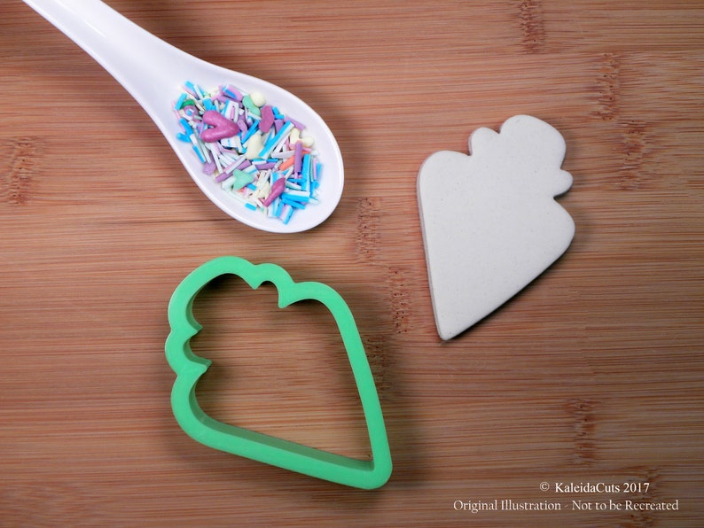 May include: A white ceramic ice cream cone cookie cutter, a green plastic ice cream cone cookie cutter, and a white ceramic ice cream cone with sprinkles in a white spoon on a wooden surface.