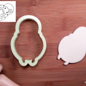 May include: A green plastic cookie cutter shaped like a girl wearing a swimsuit and a flower crown. A white cookie is shown next to the cutter, also shaped like a girl wearing a swimsuit and a flower crown.