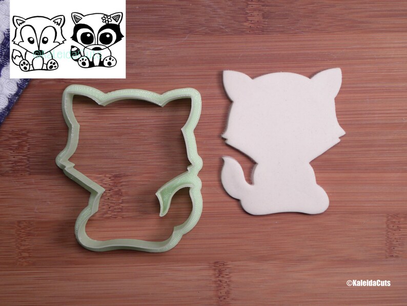 May include: A green plastic cookie cutter in the shape of a cartoon raccoon. The cutter is on a wooden surface next to a white fondant cutout of the same raccoon.