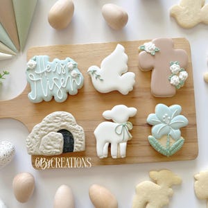 May include: Assortment of Easter-themed sugar cookies on a wooden board. Cookies include a cross, dove, lamb, flower, and a tomb with the words "He is Risen". The cookies are decorated with pastel colors and floral accents. Wooden eggs are scattered around the board.