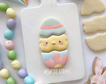 May include: A decorated sugar cookie with a chick peeking out of a cracked egg. The cookie is decorated with pastel blue, pink, and yellow icing. The cookie is on a white cutting board. The text "GBB CREATIONS" is visible on the board.