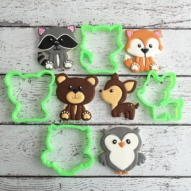 May include: A set of nine cookie cutters featuring various woodland animals, including a raccoon, fox, bear, deer, owl, and three generic animal shapes. The cutters are made of green plastic and are arranged on a white wooden surface.