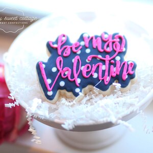 May include: A blue and white cookie decorated with pink icing and white polka dots. The icing says "be my valentine".