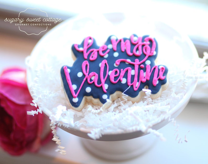May include: A blue and white cookie decorated with pink icing and white polka dots. The icing says "be my valentine".