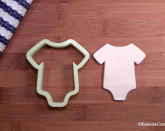May include: A green plastic cookie cutter shaped like a baby onesie, next to a white fondant onesie on a wooden surface.