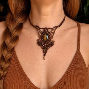 Puede incluir: Un collar de macramé marrón con una piedra preciosa ovalada de color amarillo-marrón en el centro. El collar presenta un intrincado trabajo de nudos con pequeñas cuentas doradas y un diseño de colgante. El collar se lleva puesto.
