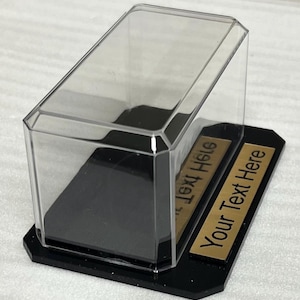 May include: A clear acrylic display case with a black base and a gold plaque that says "Your Text Here".