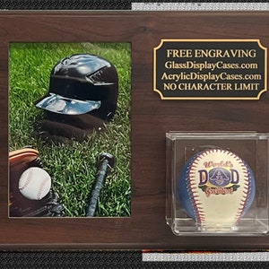 May include: A brown wooden plaque with a baseball helmet and bat in a photo frame on the left side. On the right side is a clear acrylic display case with a baseball that says "World's Greatest Dad" in gold lettering. The plaque has a gold plaque with the text "FREE ENGRAVING GlassDisplayCases.com AcrylicDisplayCases.com NO CHARACTER LIMIT".