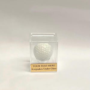May include: A white golf ball encased in a clear acrylic cube with a gold plaque that reads "YOUR TEXT HERE Keepsakes Under Glass".