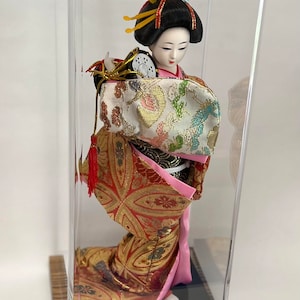 May include: A Japanese geisha doll in a clear acrylic display case. The doll wears a colorful kimono with gold accents and a traditional hairstyle. The display case sits on a wooden base with a brass plate.