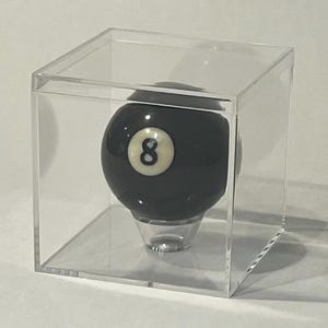 May include: A black billiard ball with a white circular inlay featuring the number eight in black, resting on a small clear stand inside a transparent, square acrylic display case that protects the ball while allowing full visibility.