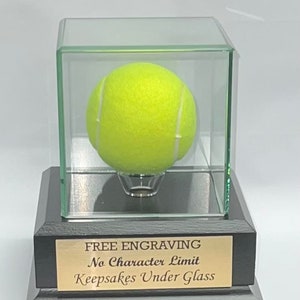 May include: A glass display case with a black base and a gold plaque that reads "FREE ENGRAVING No Character Limit Keepsakes Under Glass". Inside the case is a yellow tennis ball.
