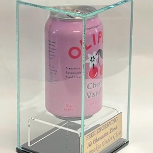 May include: A pink Olipop Cherry Vanilla soda can inside a clear glass display case. The can features text including "Prebiotics Botanicals Plant Fiber" and "Non GMO". A small gold plaque reads "FREE ENGRAVING No Character Limit Keepsakes Under Glass."