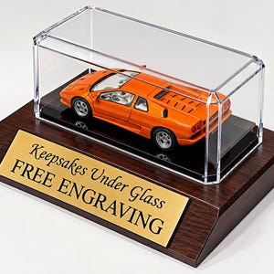May include: An orange die-cast sports car model enclosed in a clear rectangular acrylic display case, mounted on a dark brown wooden base with a gold-colored metal plaque featuring the black text Keepsakes Under Glass FREE ENGRAVING.