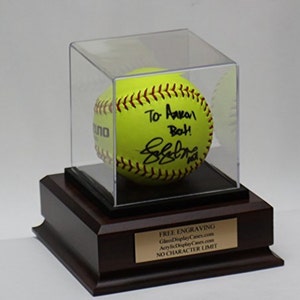 May include: A yellow softball with red stitching is displayed in a clear acrylic case on a wooden base. The softball is signed "To Avaan Both!" with a signature. The base has a gold plaque that reads "FREE ENGRAVING GlassDisplayCases.com AcrylicDisplayCases.com NO CHARACTER LIMIT"