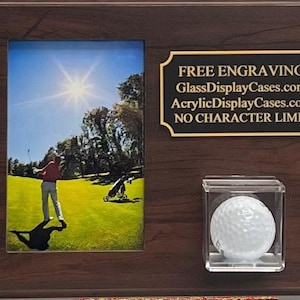 May include: A wooden display case with a golf theme. It has a framed photo of a golfer on a green, a clear acrylic case holding a golf ball, and a plaque with text. The plaque reads "FREE ENGRAVING" and website addresses.