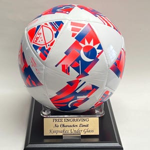 May include: A white soccer ball with red and blue geometric patterns and the MLS logo, resting on a clear acrylic stand atop a black wooden base. A gold-colored plaque on the base features the text: FREE ENGRAVING, No Character Limit, Keepsakes Under Glass.