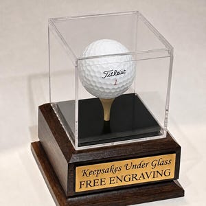 May include: A white Titleist golf ball with the number 1 sits on a wooden tee inside a clear acrylic display case. The case rests on a dark brown wooden base featuring a gold-colored metal plaque with the text Keepsakes Under Glass FREE ENGRAVING.