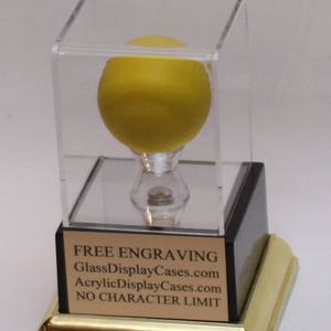 May include: A yellow sphere on a clear acrylic base inside a clear acrylic cube with a black base and a gold trim. The base has the text "FREE ENGRAVING GlassDisplayCases.com AcrylicDisplayCases.com NO CHARACTER LIMIT".