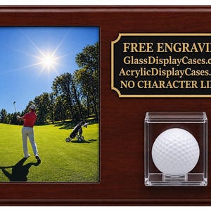 May include: A dark wood display plaque featuring a vertical photo frame with a golfer on a green course, a clear acrylic cube containing a white golf ball, and a black plate with gold text: FREE ENGRAVING GlassDisplayCases.com AcrylicDisplayCases.com NO CHARACTER LIMIT.