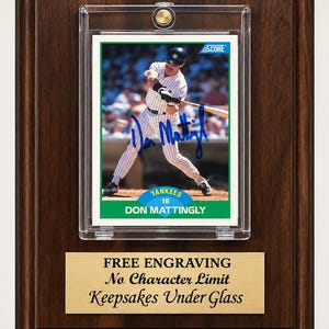 May include: A dark wood plaque featuring a Don Mattingly baseball card in a clear protective case. The card shows a player in a pinstriped uniform with a blue ink autograph. Below the card is a gold-toned metal plate with the text: FREE ENGRAVING, No Character Limit, Keepsakes Under Glass.