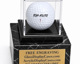 May include: A white Top-Flite golf ball with the number 1 printed on it, enclosed in a clear acrylic display case. The case sits on a black faux marble base featuring a gold-colored metal plaque with the text FREE ENGRAVING GlassDisplayCases.com AcrylicDisplayCases.com NO CHARACTER LIMIT.