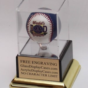 May include: A clear acrylic display case with a black base and gold trim. Inside the case is a white baseball with blue and red stitching. The baseball has the words "World's Greatest Dad" printed on it.